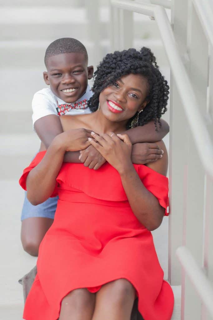 Black woman wearing a red dress with her young son hugging her from behind.