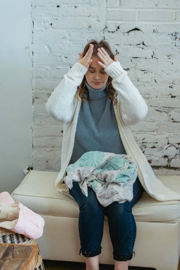 Woman sitting with clothes on lap holding her head looking stressed.
