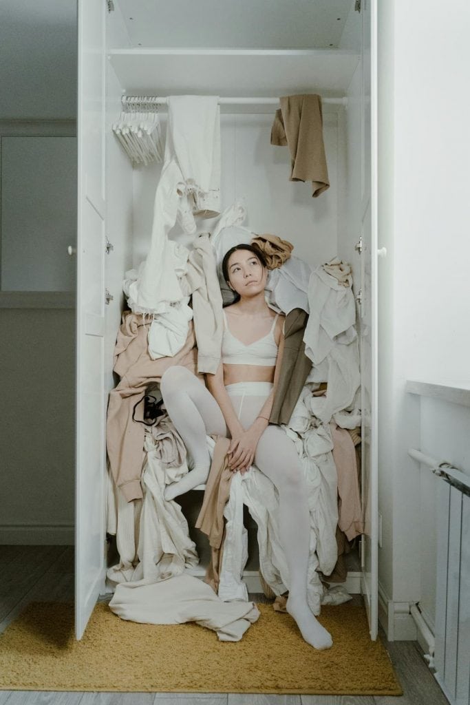 Woman in a bra and tights sitting in a messy closet full of clothes that aren't hung up.