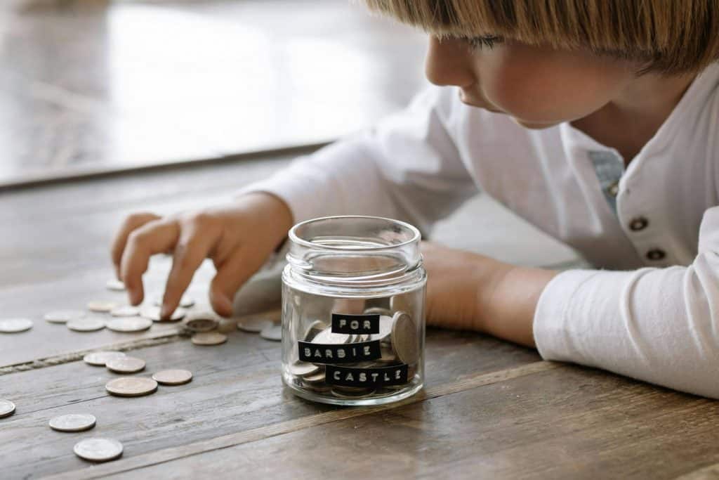 Kid putting coins into a clear jar that says "for barbie castle" on it.