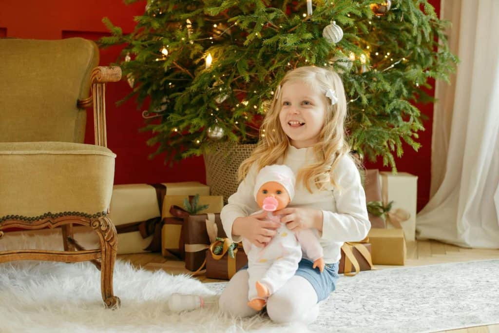 Girl holding a doll in front of a Christmas tree and smiling. 