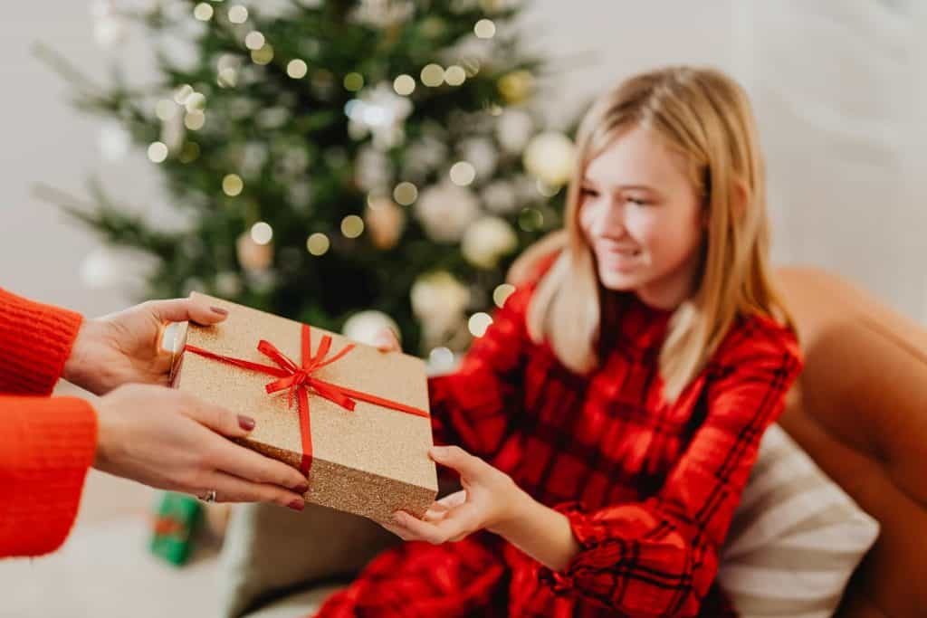 Older female child being handed a gift with a Christmas tree in the background. 