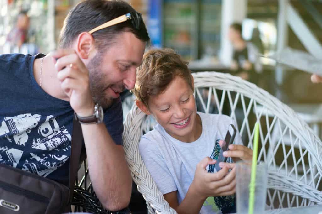 Kid sitting next to his dad smiling while looking at a kid budgeting app on his phone. 