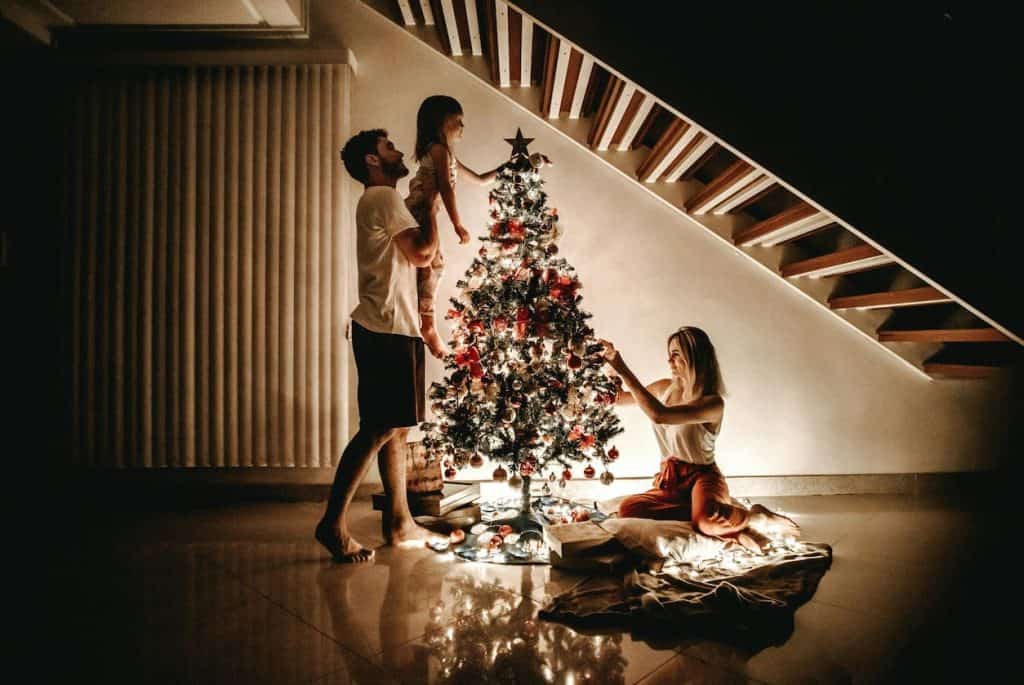 Dad holding daughter up to put star on tree while mom is sitting on the floor fixing the ornaments. 