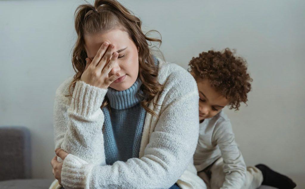 A very exhausted and overwhelmed mom sitting with her hand on her forehead. Her son is sitting behind her. 