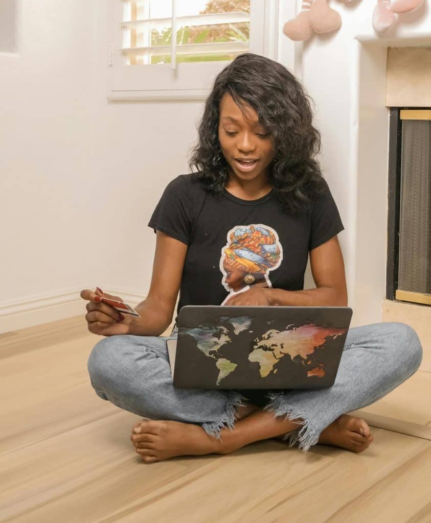 Woman in a black shirt sitting crossed legged on floor with computer in lap looking at her credit card. 