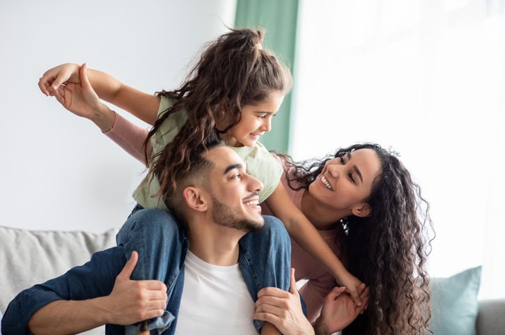 Young girl sitting on dad's shoulders while smiling and looking at her mom. 