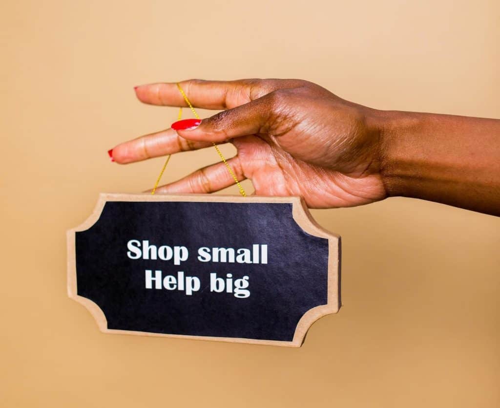 Hand with red nail polish holding a small sign that says shop small, help big. 
