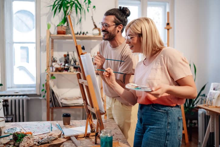 Guy with a man bun and woman with blond hair each painting on a canvas. 