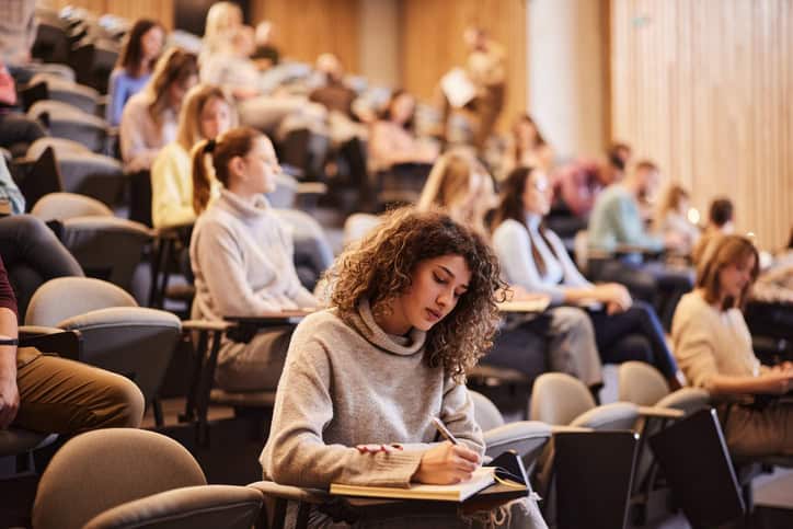 Young women with curly hair sitting in a lecture hall at a university taking notes. 