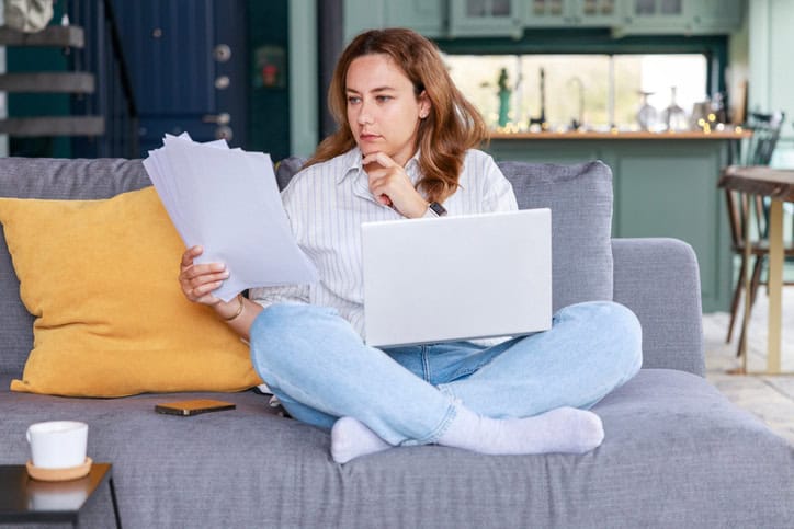 Women sitting on couch crossed legged looking at papers with a laptop on her lap. 