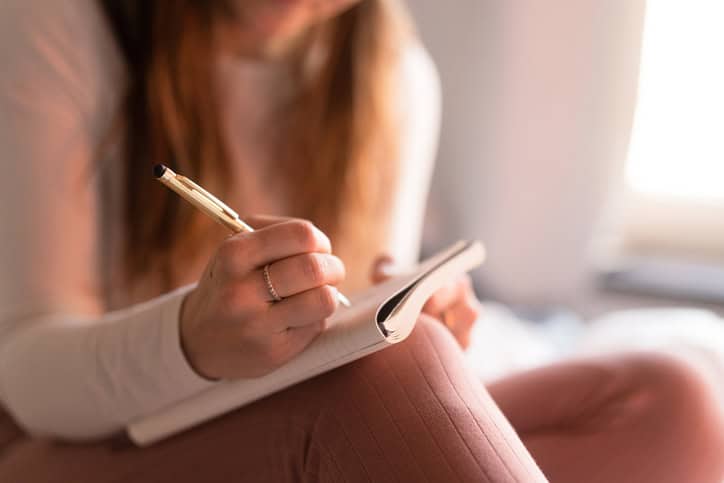 Image of a woman's hands holding a notebook on her knee with a pen in hand. 