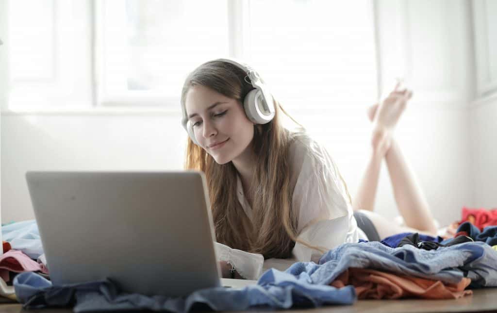 Woman laying on her stomach with feet crossed looking at lap top with headphones on. 