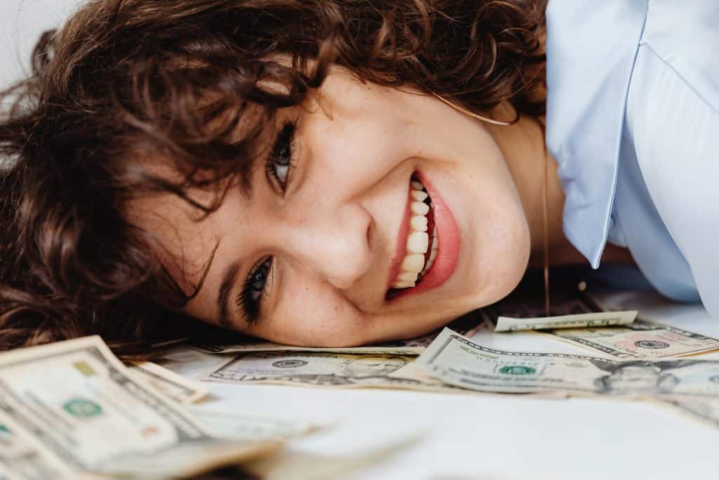 A woman with an abundance money mindset laying her head on a pile of money with a big smile on her face.