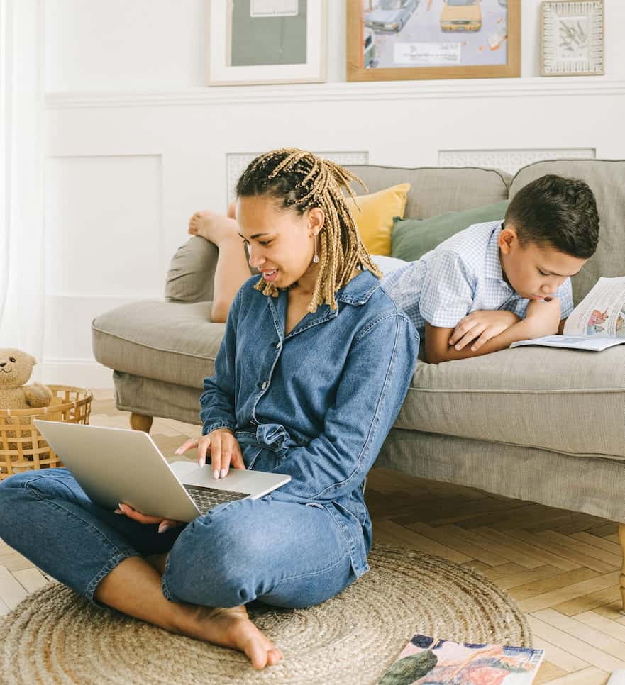 Mom with a growth money mindset sitting on the floor in front of the couch. She is on her laptop with her son laying on the couch behind her reading a book.