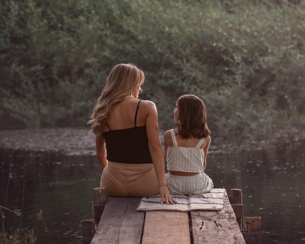 The backs of a mom and daughter sitting on a dock looking at each other.