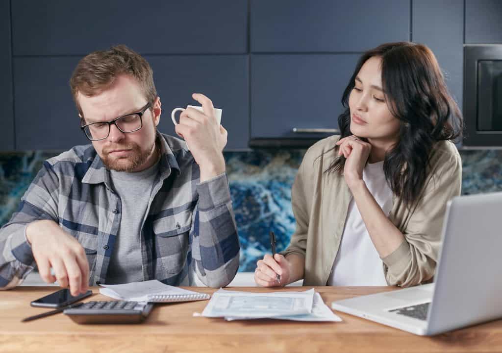 A man and a woman sitting at the table with a calculator and laptop looking stressed about money.