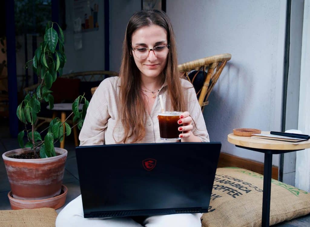 A woman wearing glasses who is sitting on her laptop working on her eBay business with a drink in her hand. 