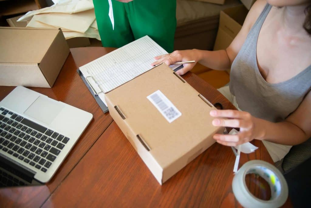 Shows a woman's hands holding a small shipping box with a list next to her to track eBay sales. 