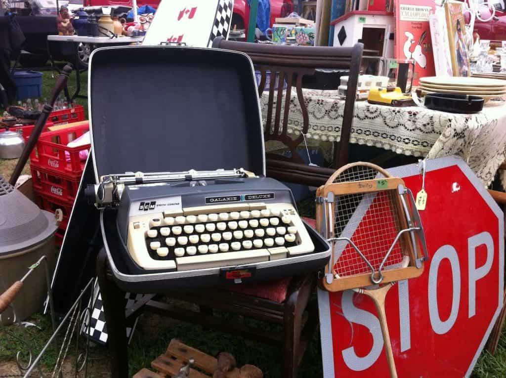 Shows an old typewrite, stop sign, and other items at what looks to be a flea market or yard sale. 