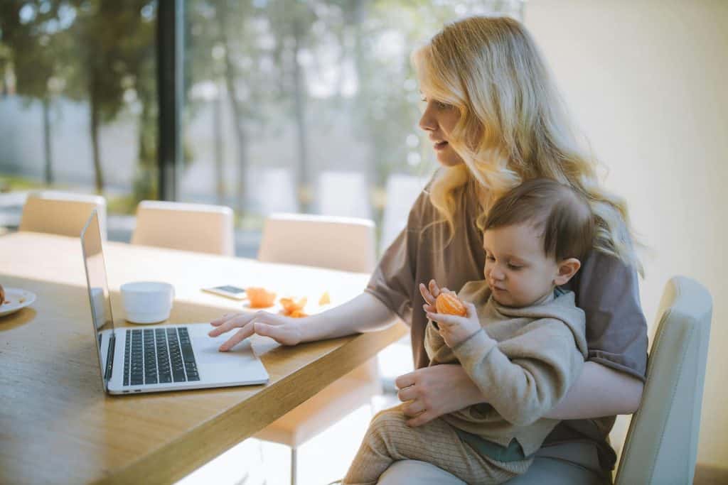 Woman with blond hair sitting in a chair with her son on her lap while she is working on her laptop tracking her eBay expenses and profits. 