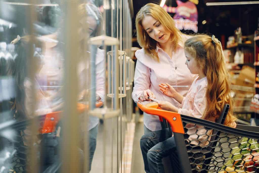 Image of a mom and daughter shopping. The girl is sitting in the cart with a grocery list and pen in hand. 