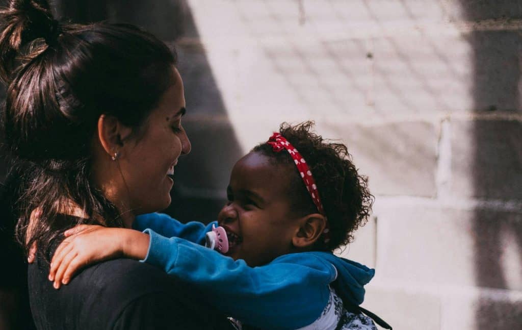 An image of a mom holding her daughter. They are both looking at each other with big smiles on their faces. The daughter has a pacifier. 